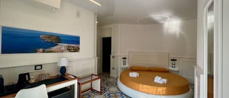 Habitación de hotel con cama, escritorio, silla, una mesita, lámpara y un cuadro de un paisaje costero.