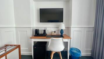 Habitación de hotel con un escritorio de madera, una silla blanca, un televisor montado en la pared, cafetera, lámpara, cesto de basura y un refrigerador pequeño.