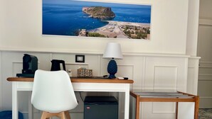 A hotel room with a desk, chair, and a small table with a lamp. There is a large window with a view of a coastal landscape. - Tea Luxury (Praia a Mare)