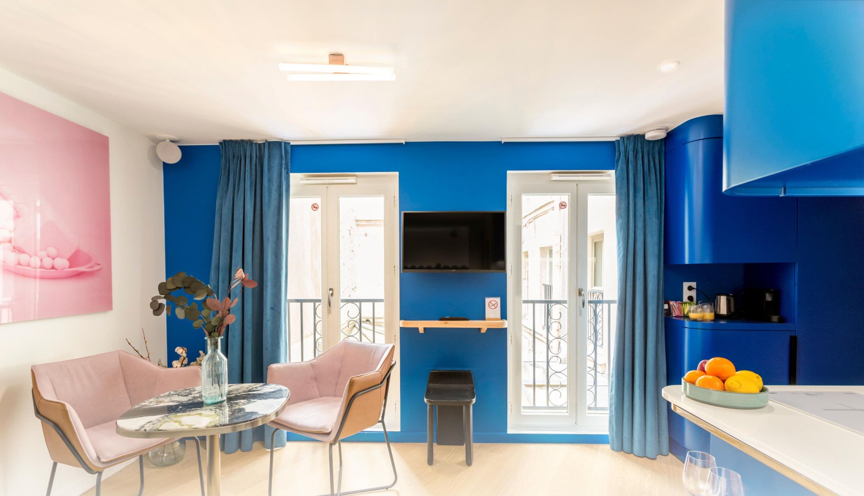 Une pièce moderne avec un mur d’accent bleu, un coin repas avec des chaises roses, une petite table agrémentée d’un vase, et un espace cuisine avec un panier de fruits.