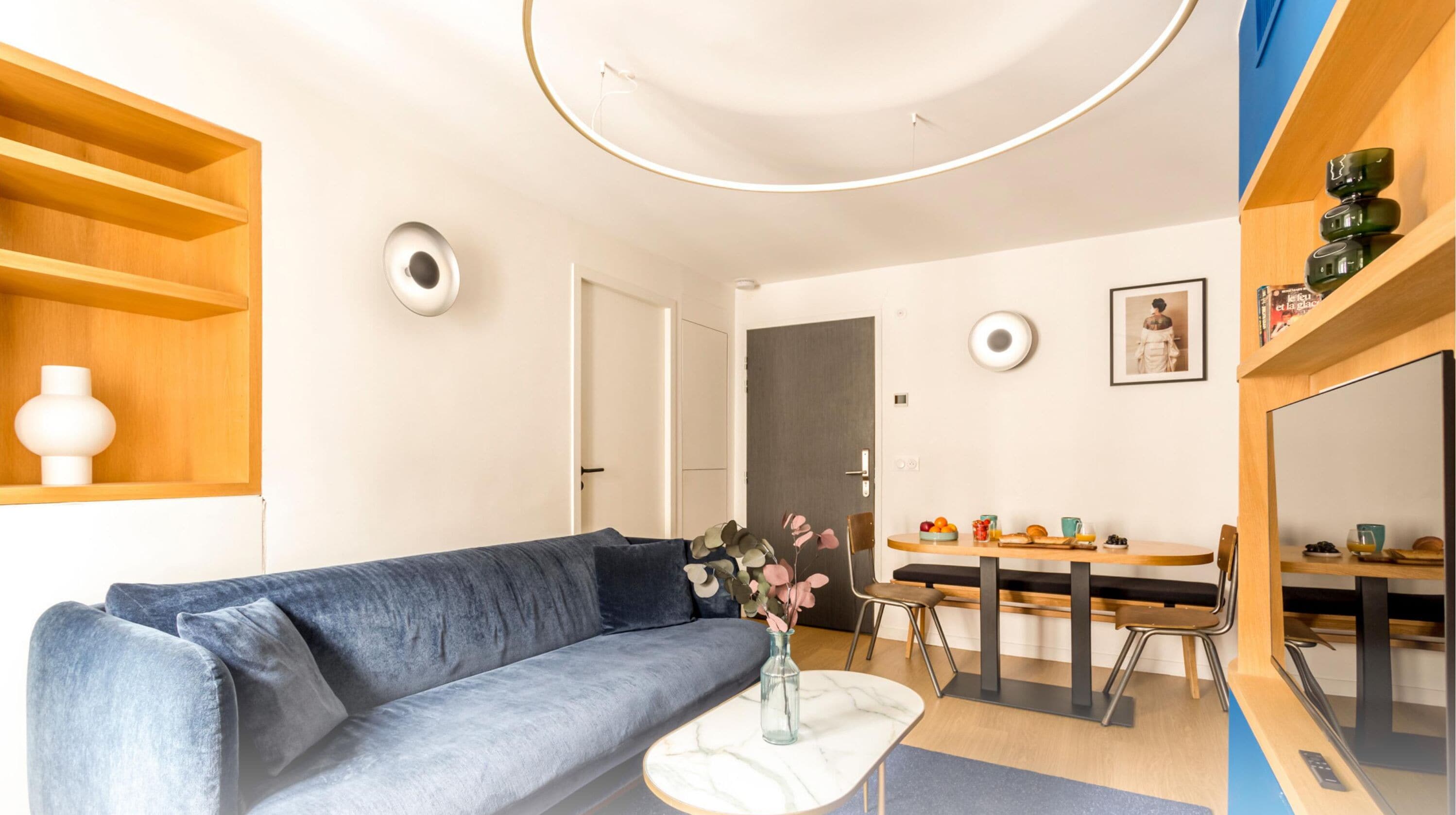 A modern living room with a blue sofa, a small white table, and a dining area with a wooden table and chairs.