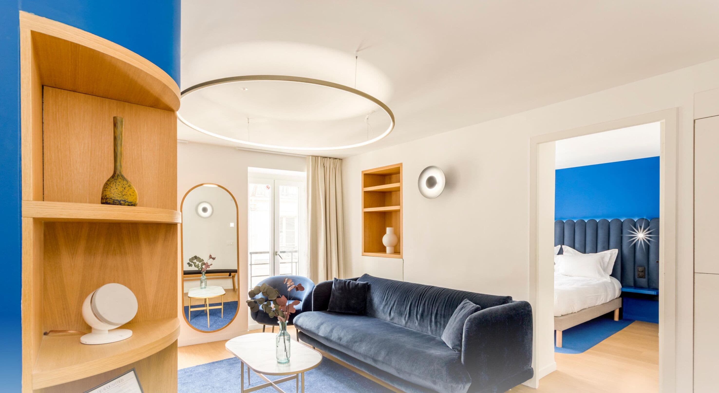 Una habitación de hotel moderna con un sofá gris, un espejo redondo, un estante de madera con objetos decorativos, una mesita con un florero y una cama con cabecera azul.