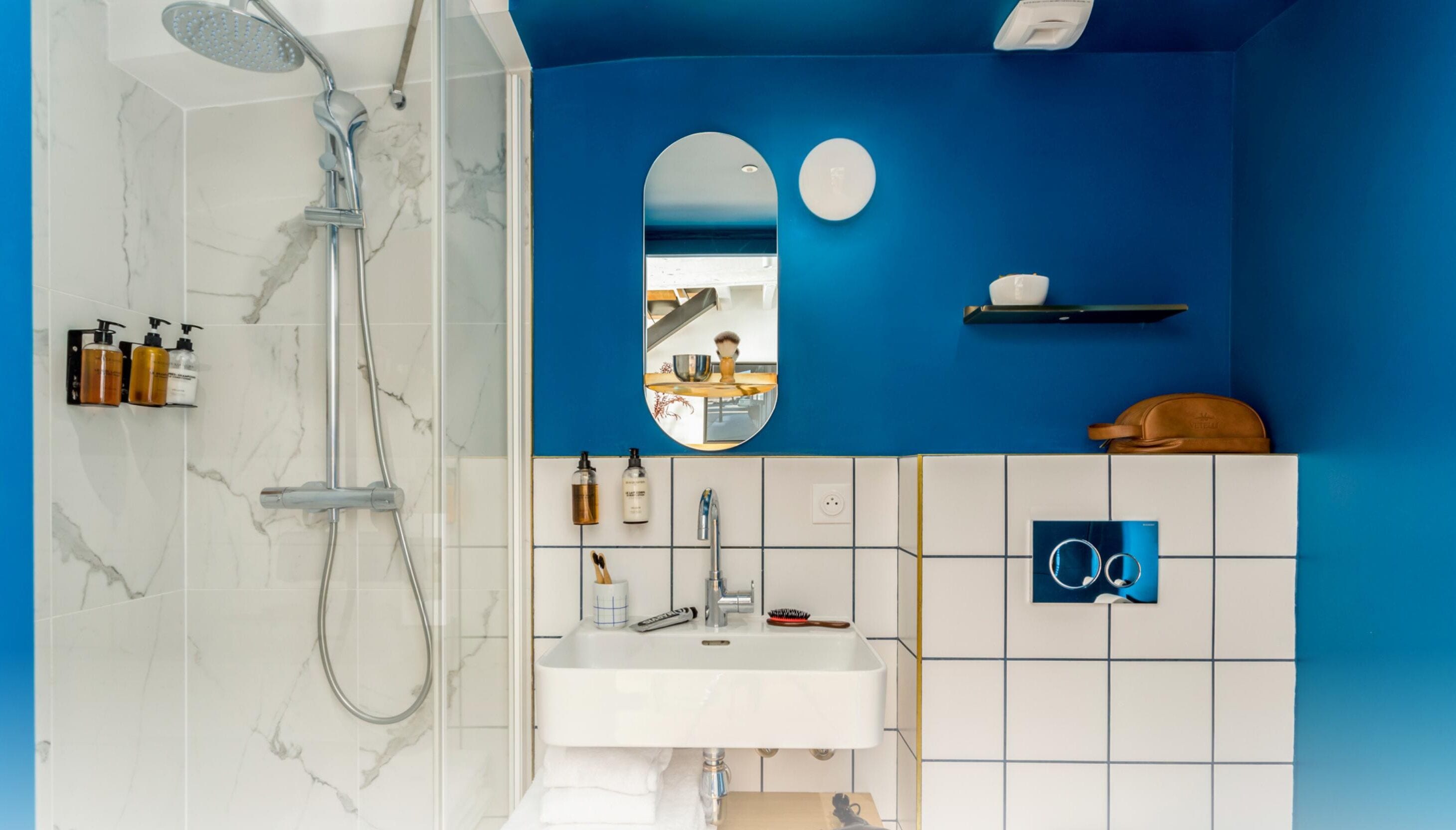 Baño con ducha, lavamanos y espejo. Hay artículos de aseo en la pared y un perchero para toallas.
