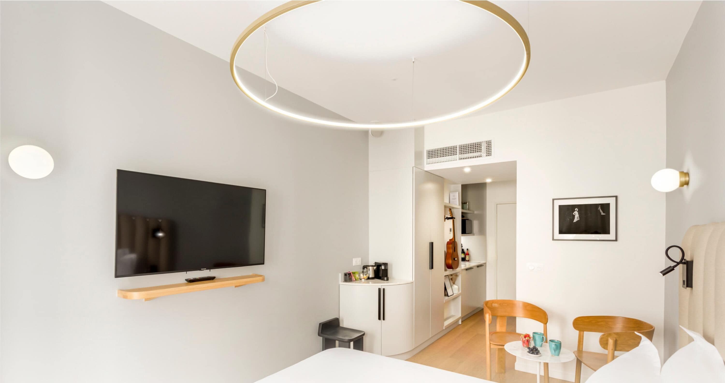 A modern hotel room with a flat-screen TV, a wooden TV stand, a dining area with a small table and chairs, and a wardrobe with built-in shelves.