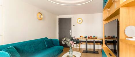 A modern living room with a teal sofa, a dining area with a wooden table, and a TV on the right.
