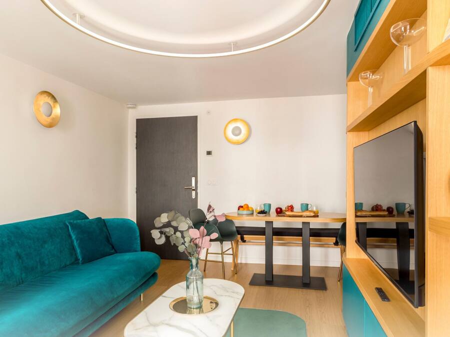 A modern living room with a teal sofa, a dining area with a wooden table, and a TV on the right.