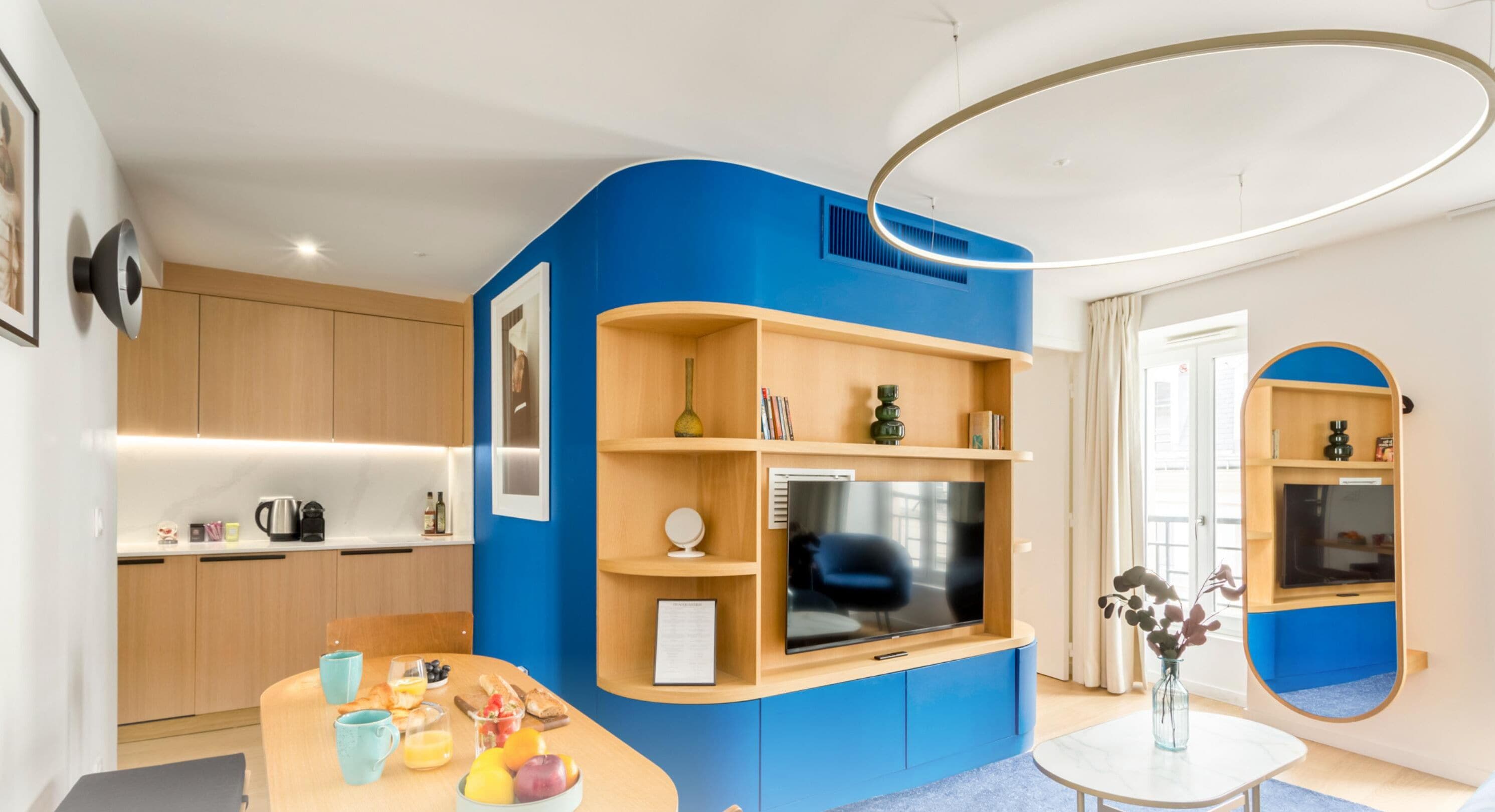 Un salon moderne avec un mur d’accent bleu, un téléviseur à écran plat, une machine à café et un coin repas avec une table dressée pour deux personnes.