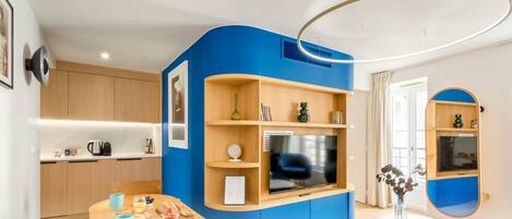 Un salon moderne avec un mur d’accent bleu, un téléviseur à écran plat, une machine à café et un coin repas avec une table dressée pour deux personnes.