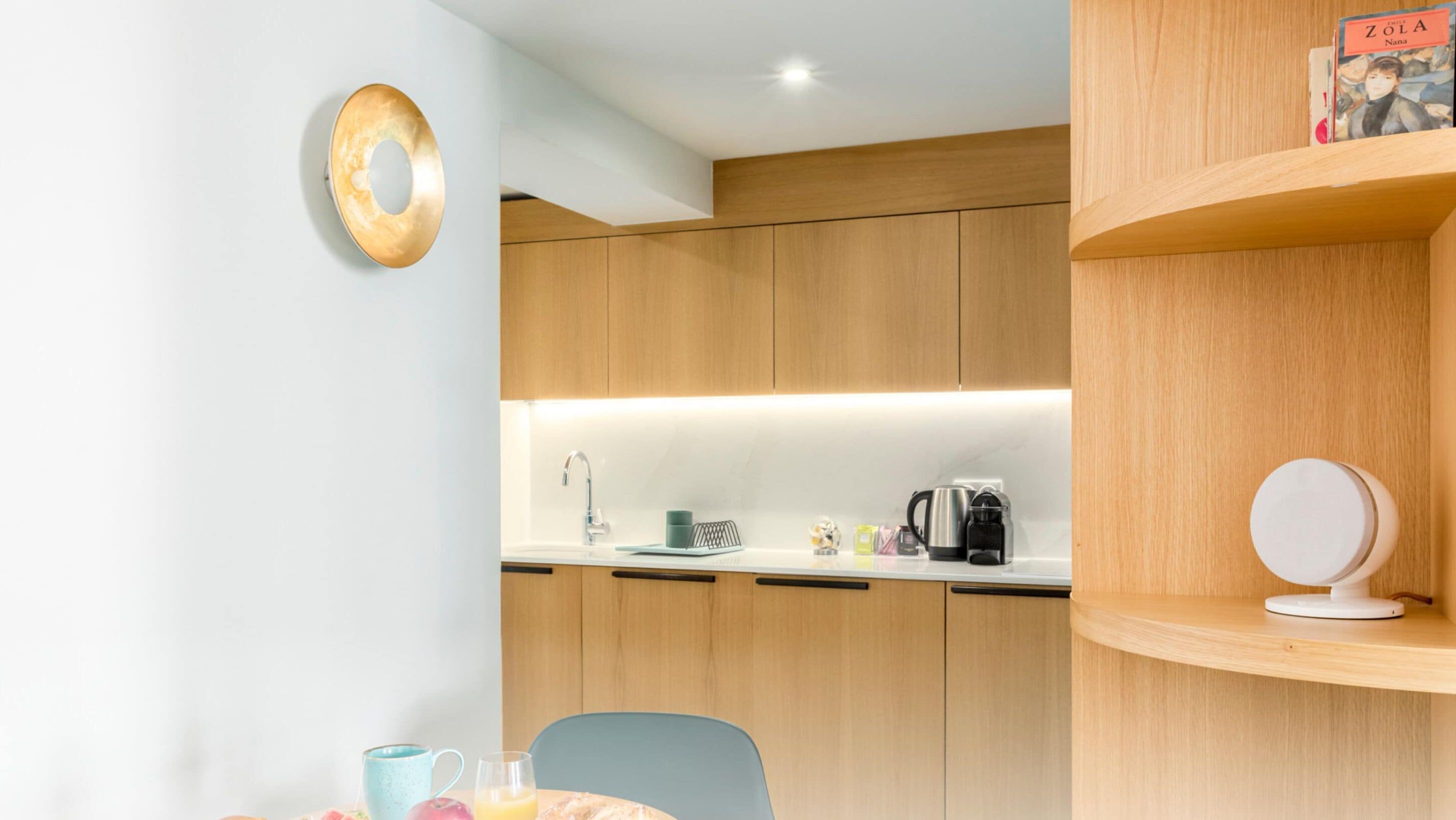 A modern kitchen with wooden cabinets, a countertop with a kettle, coffee maker, and a book titled "Zola Nana".