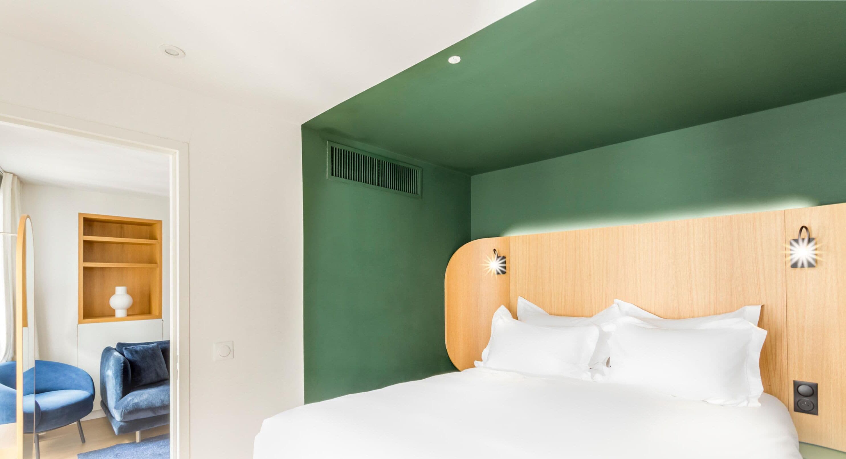 A hotel room with a large bed, a wooden headboard, a small shelf with a vase, and a blue chair.