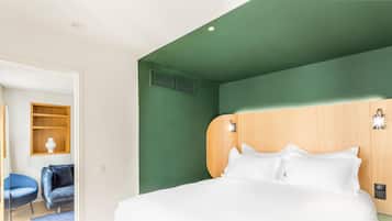 A hotel room with a large bed, a wooden headboard, a small shelf with a vase, and a blue chair.