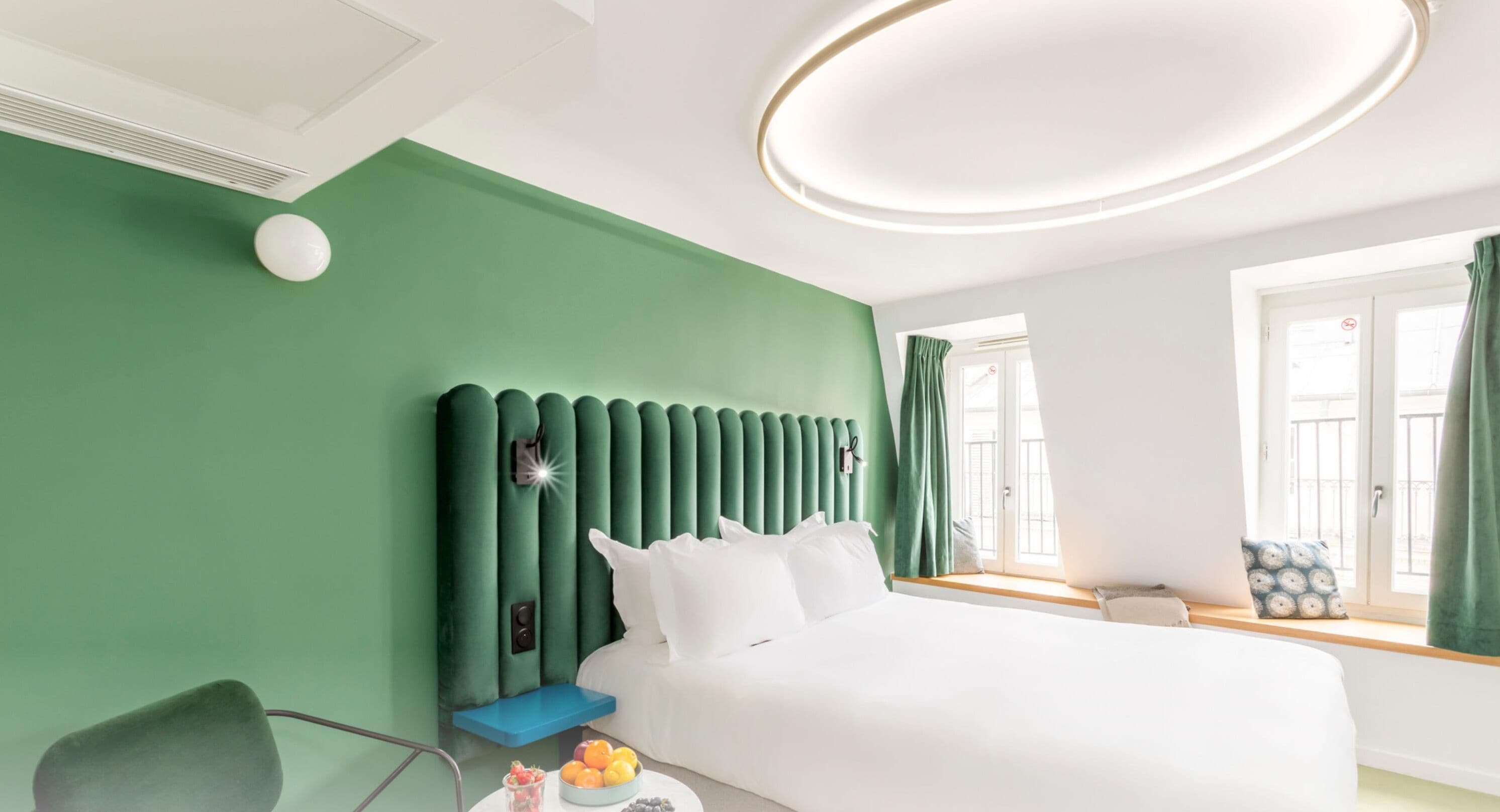 A modern hotel room with a green accent wall, a large bed with white bedding, a bedside table with fruit, and a window with curtains.