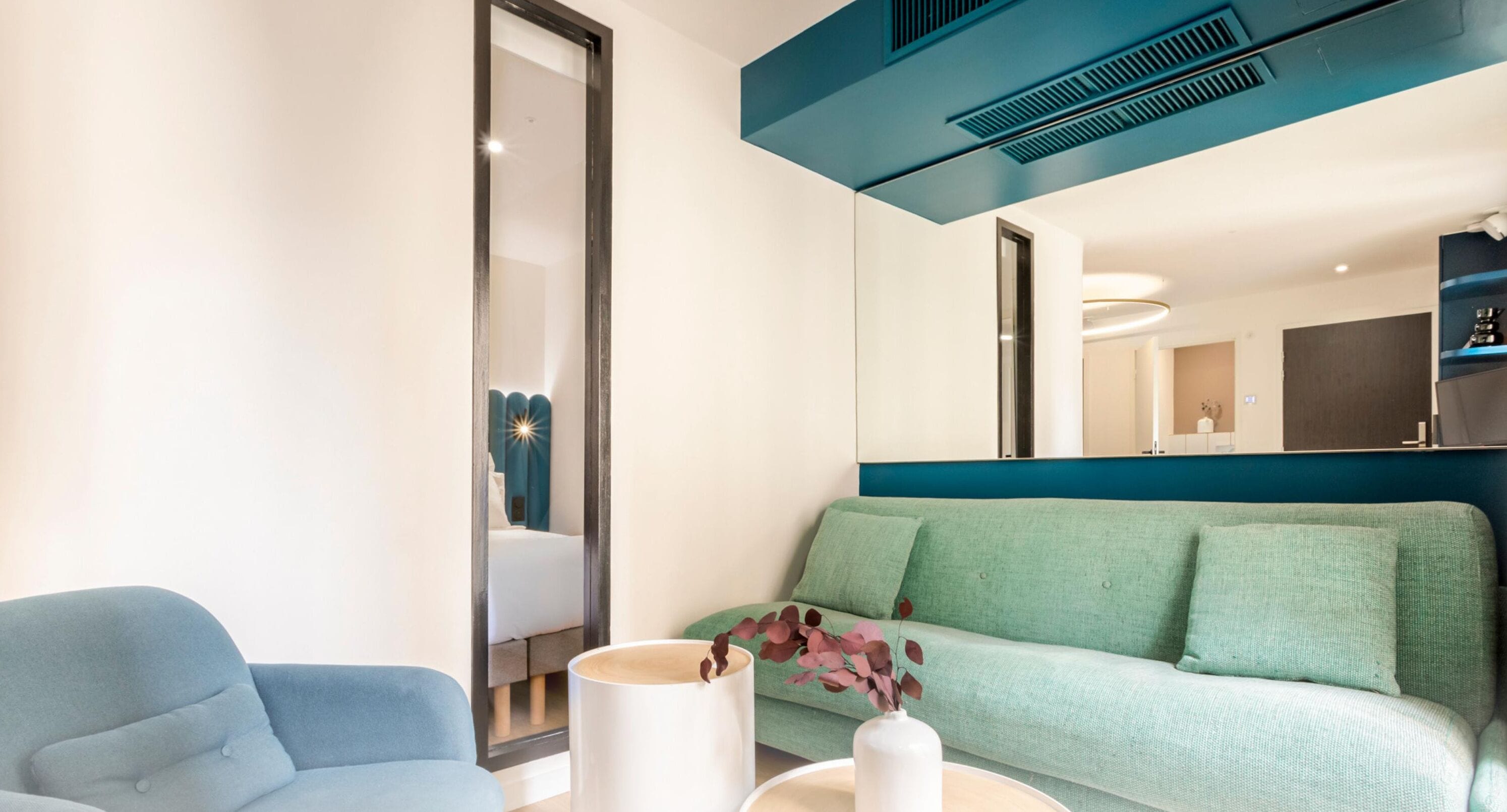 Una habitación de hotel moderna con un sofá azul, una mesita auxiliar blanca con un florero y un espejo grande que refleja una cama y un baño.