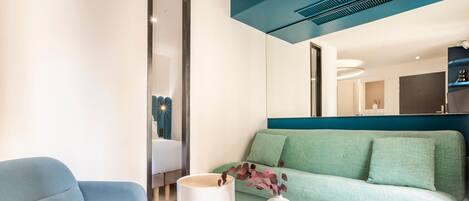 Una habitación de hotel moderna con un sofá azul, una mesita auxiliar blanca con un florero y un espejo grande que refleja una cama y un baño.