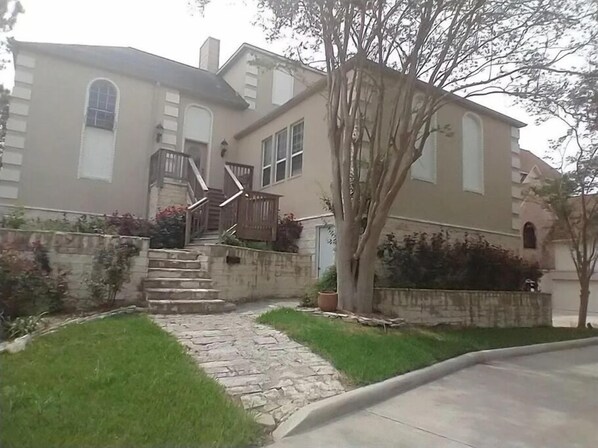 A two-story house with a stone staircase leading to the front door, surrounded by a small garden and a tree.