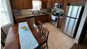 Una cocina con armarios de madera, electrodomésticos de acero inoxidable, una mesa de comedor con sillas y un ventanal con persianas.