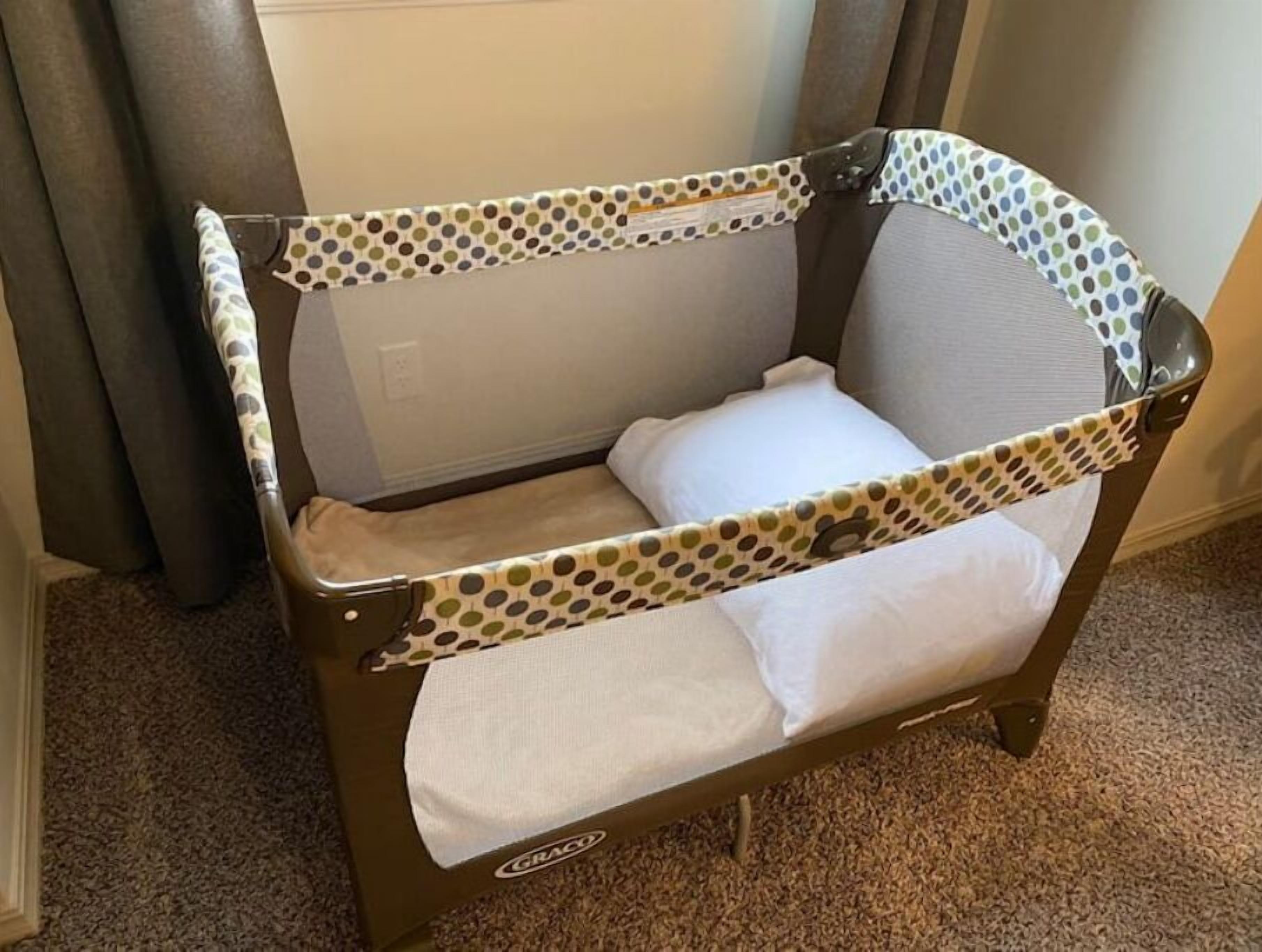 En Graco-babysäng med ett mönster av prickar, placerad mot en vägg med en gardin.