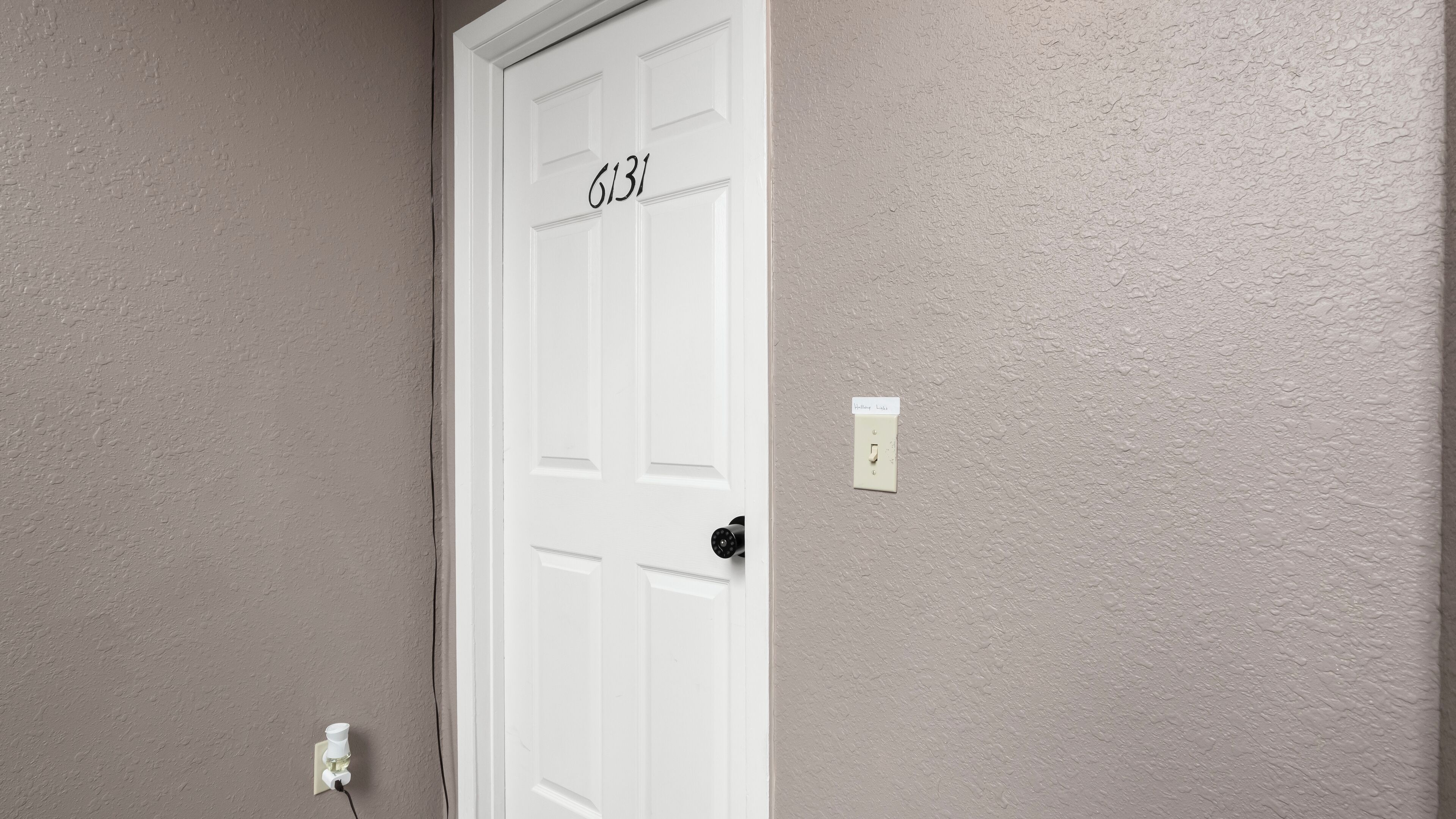 A white door with the number 6131, a wall-mounted light switch, and a power outlet with a plug.