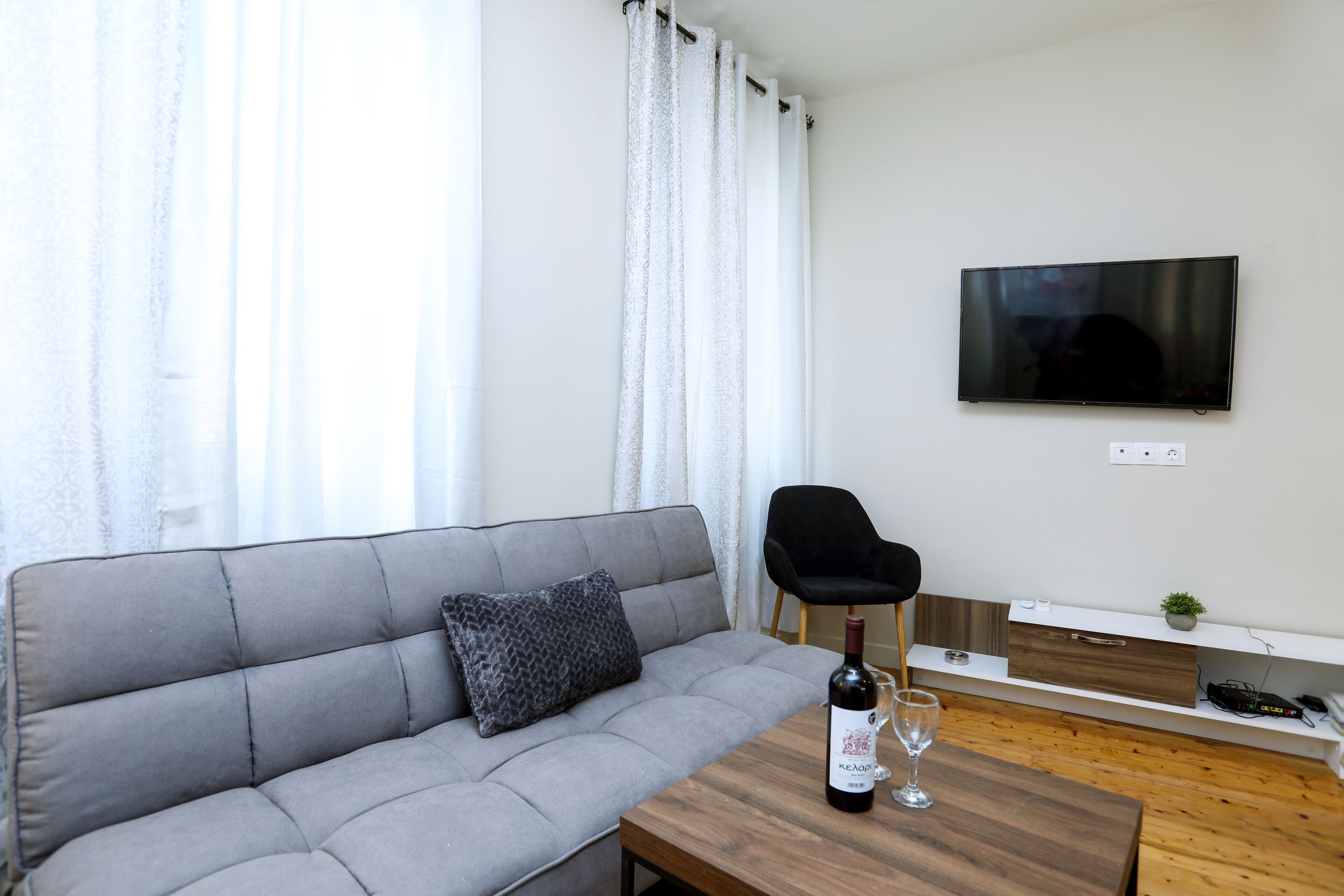 Ein modernes Wohnzimmer mit einem grauen Sofa, einem Holztisch mit einer Weinflasche und einem Glas, einem schwarzen Sessel und einem flachbildfernseher an der Wand.