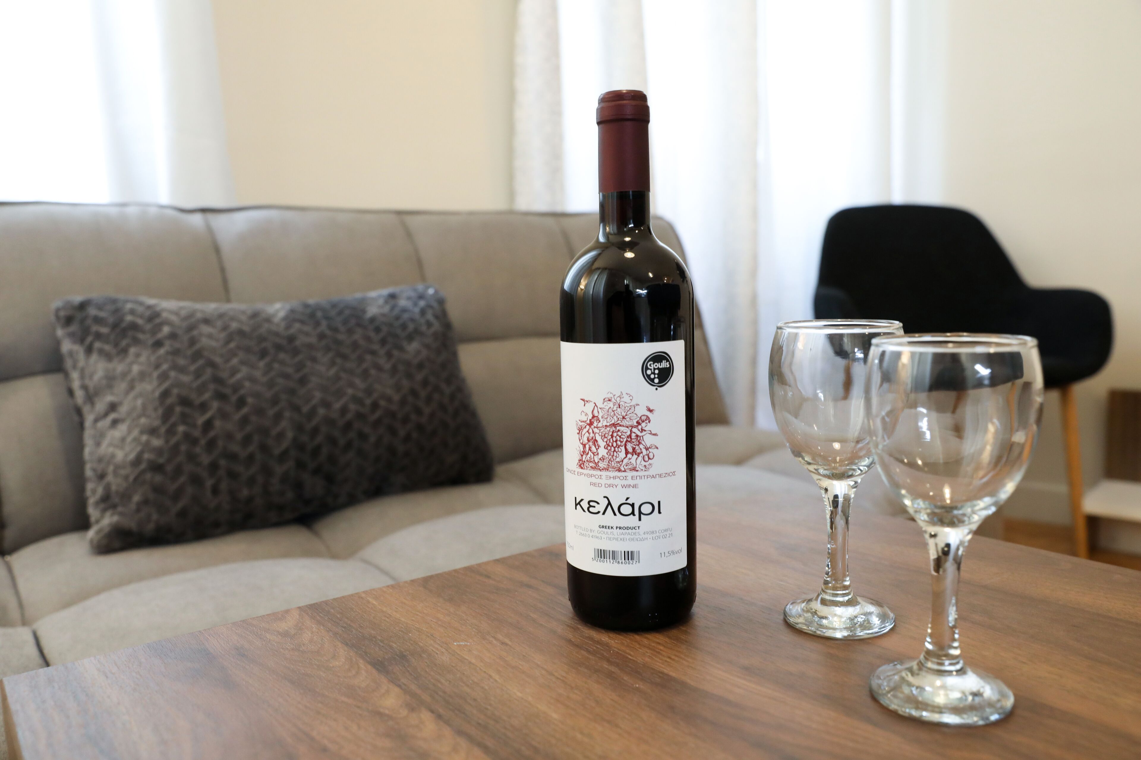 Een fles Keaapi wijn op een houten tafel, met twee wijnglazen ernaast, in een woonkamer met een bank en een zwarte stoel.