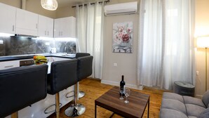 A modern kitchen with white cabinets, a stainless steel refrigerator, and a fruit bowl on the counter. There is a dining area with two black chairs and a wooden table with a wine bottle and glasses. A gray sofa and a lamp are in the living area.