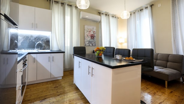 A modern kitchen with white cabinets, a black countertop, and a central island with seating. There is a window with curtains, an air conditioning unit, and a hanging light fixture.
