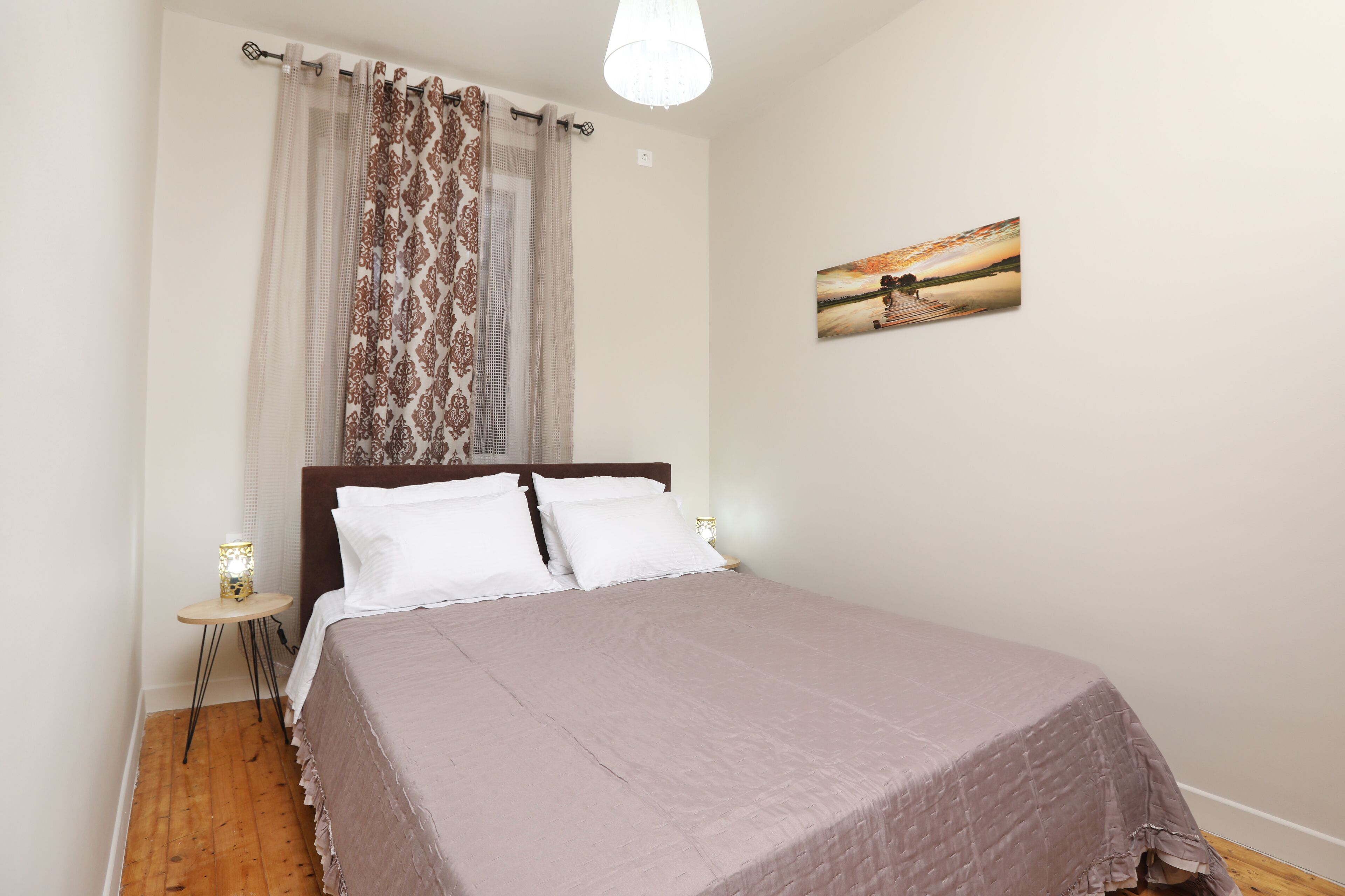 Quarto com cama, cortinas, mesa de cabeceira com luminária e um quadro na parede.