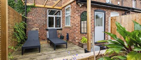 A patio with two black lounge chairs, a wooden pergola, and a brick house with a white door.