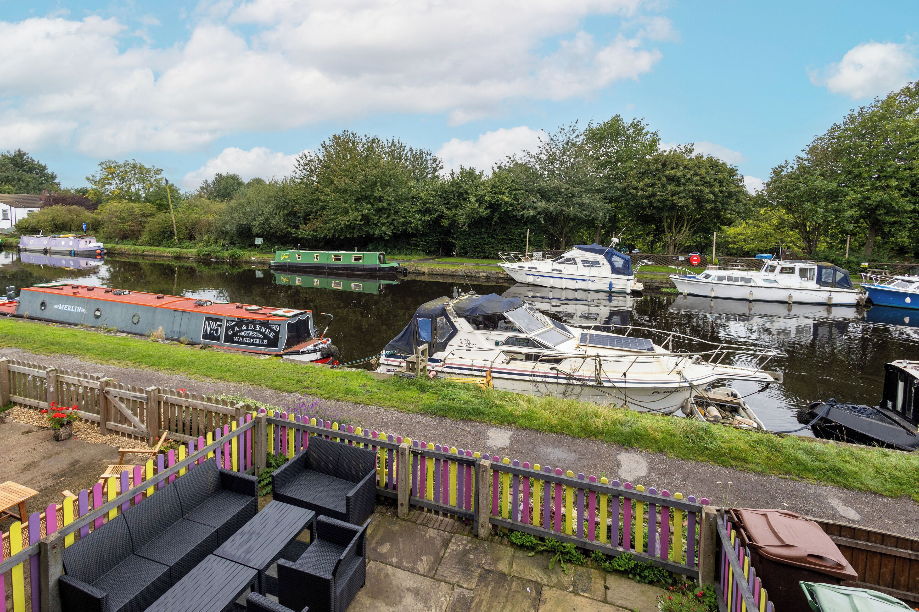 A riverside dock with various boats, a wooden fence, and outdoor seating.
