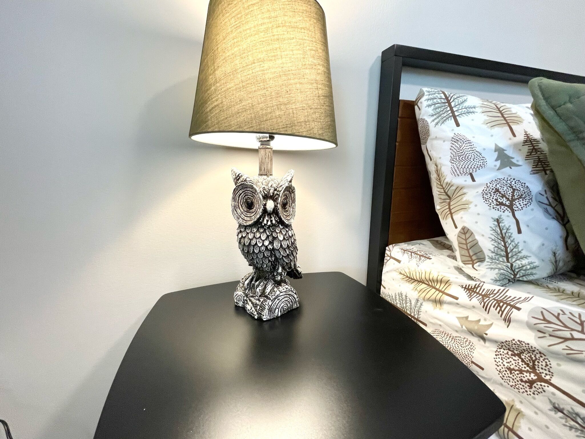 A table lamp with an owl design on a black table next to a bed with patterned bedding.