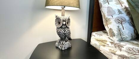 A table lamp with an owl design on a black table next to a bed with patterned bedding.
