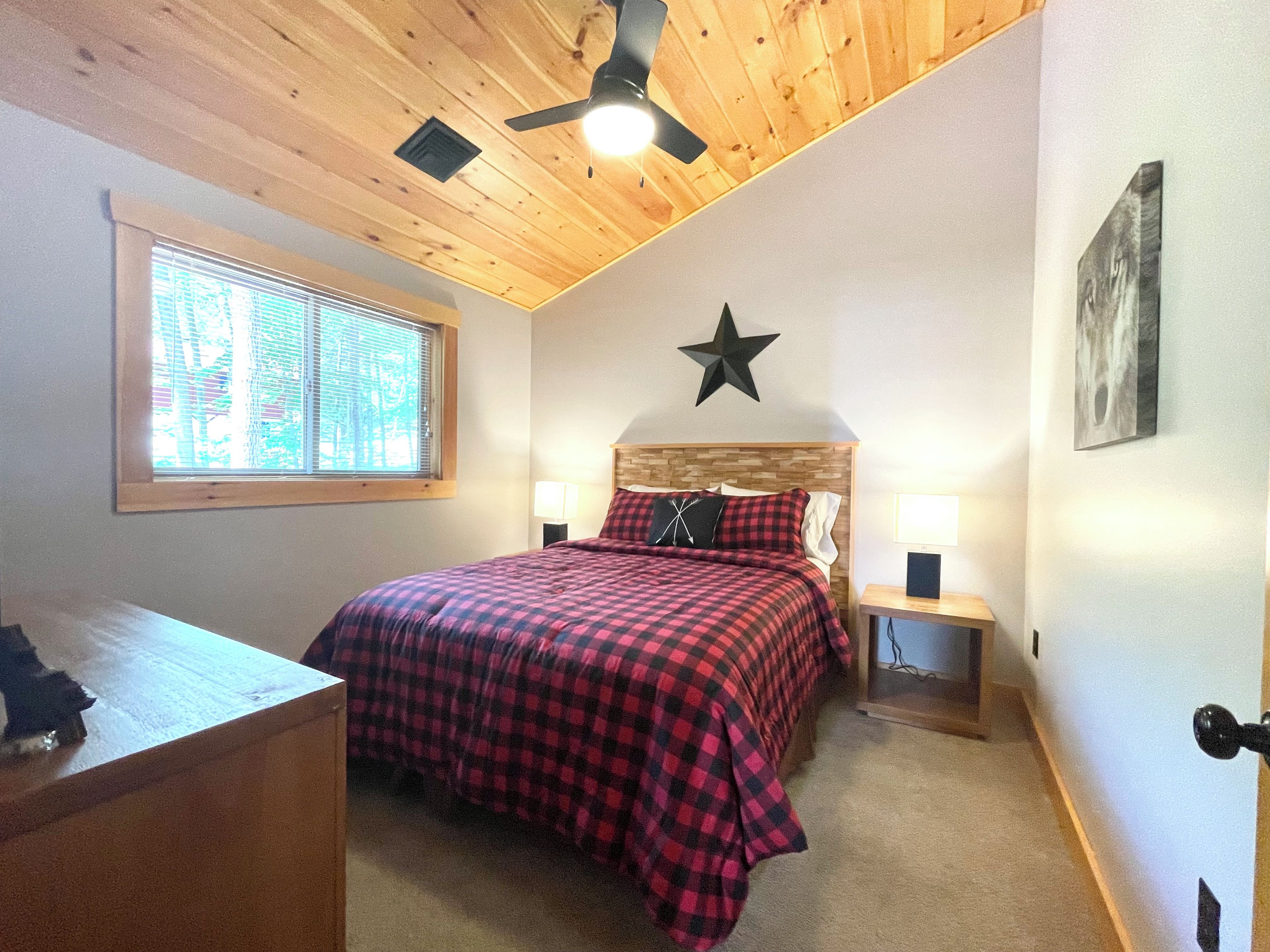 A bedroom with a wooden ceiling, a star decoration, a bed with a red and black checkered bedspread, two bedside lamps, and a window with blinds.