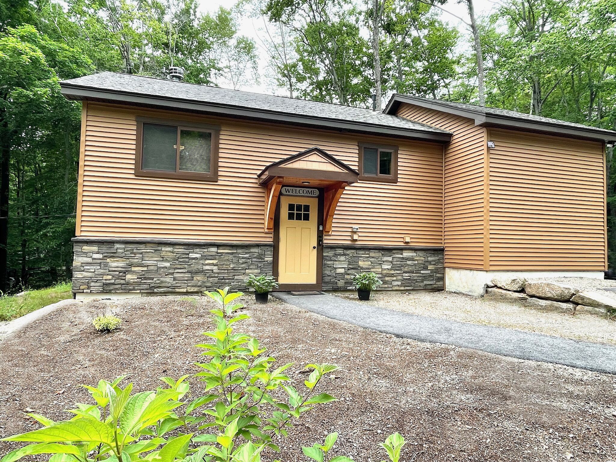 A single-story house with a stone foundation, a yellow door, and a welcome sign.