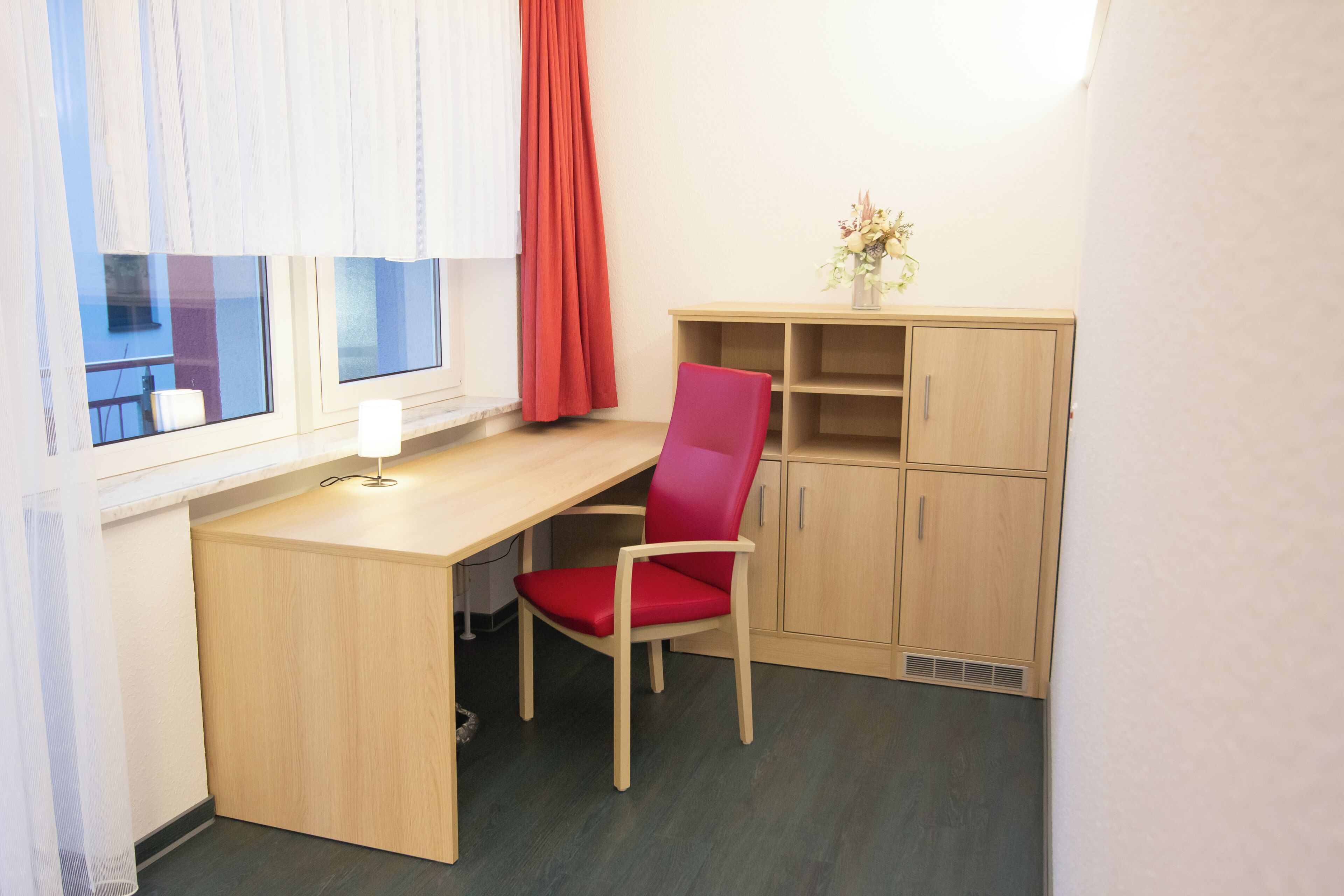 Una habitación pequeña y funcional con escritorio, silla, ventana con cortinas y un armario.