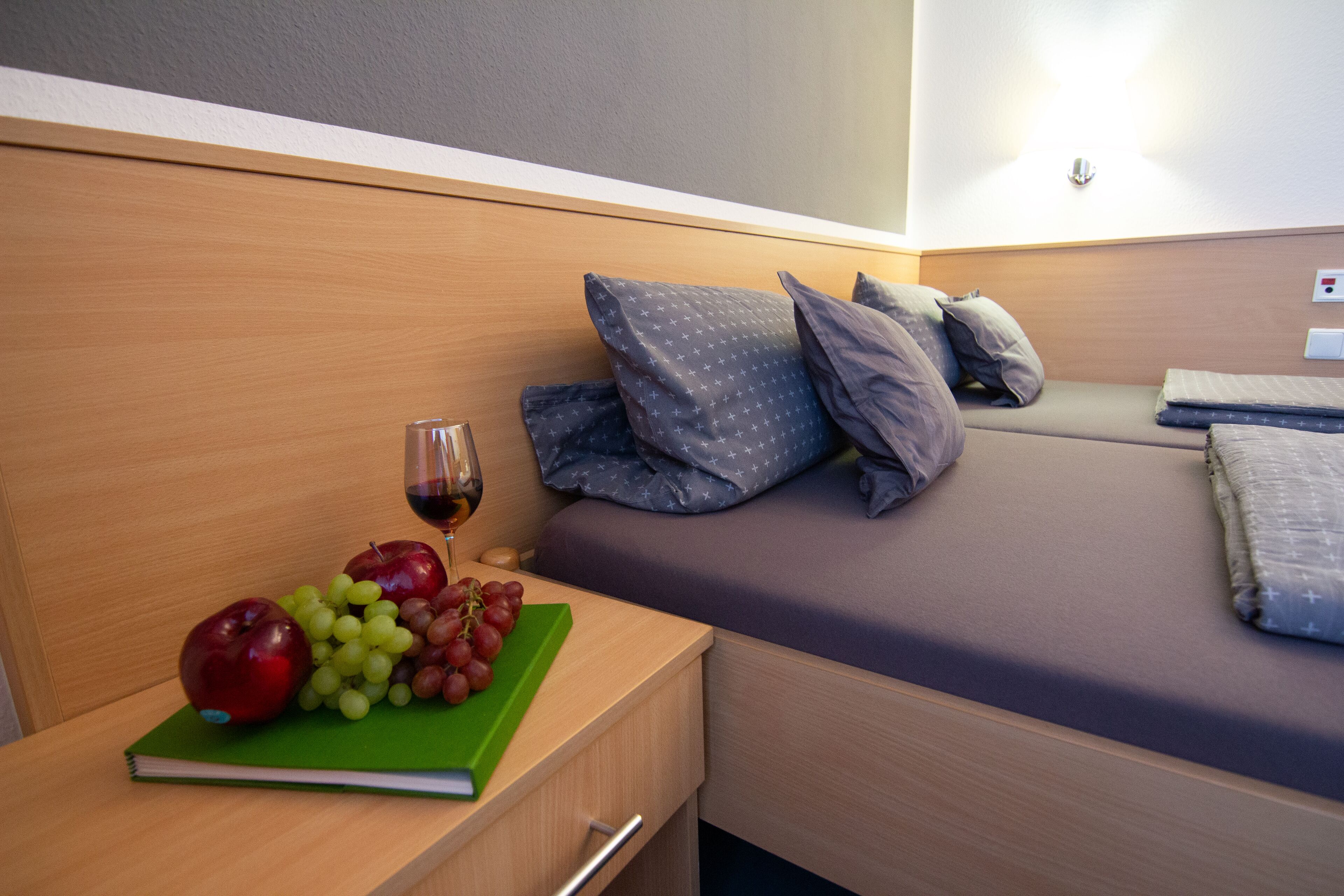 Ein ordentlich gemachtes Bett mit Kissen und einem Nachttisch mit Buch, Trauben und einem Apfel.