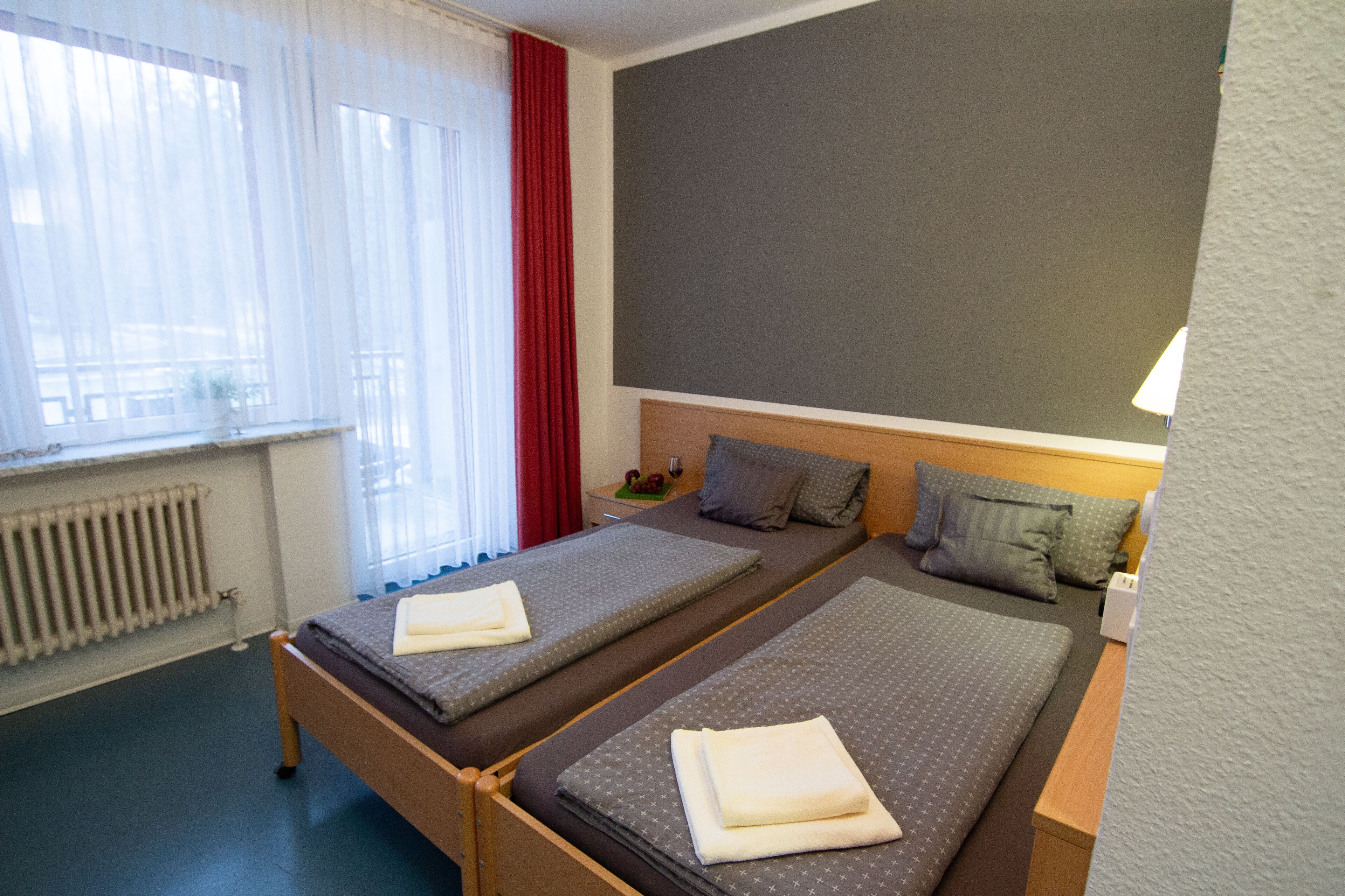 A hotel room with two beds, a radiator, a window with curtains, and a wall-mounted lamp.