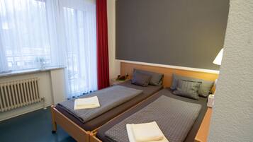 A hotel room with two beds, a radiator, a window with curtains, and a wall-mounted lamp.