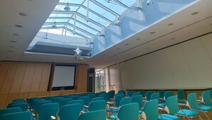 A conference room with a high ceiling and skylight, rows of teal chairs, and a projector screen. - Green Night Hotel (Falkenstein/Vogtl.)