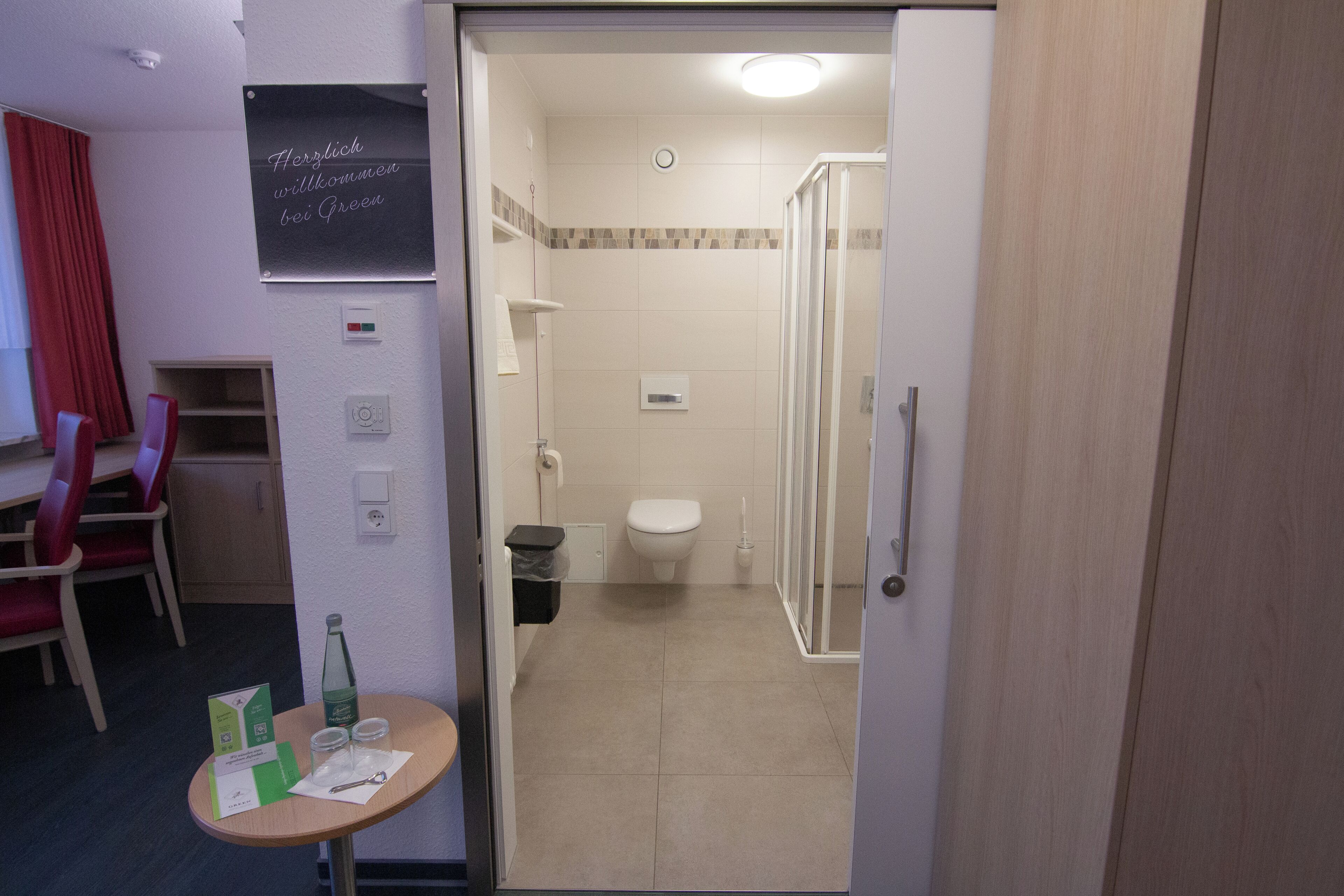 Un baño de hotel con ducha, inodoro y una mesita con una botella y vasos.