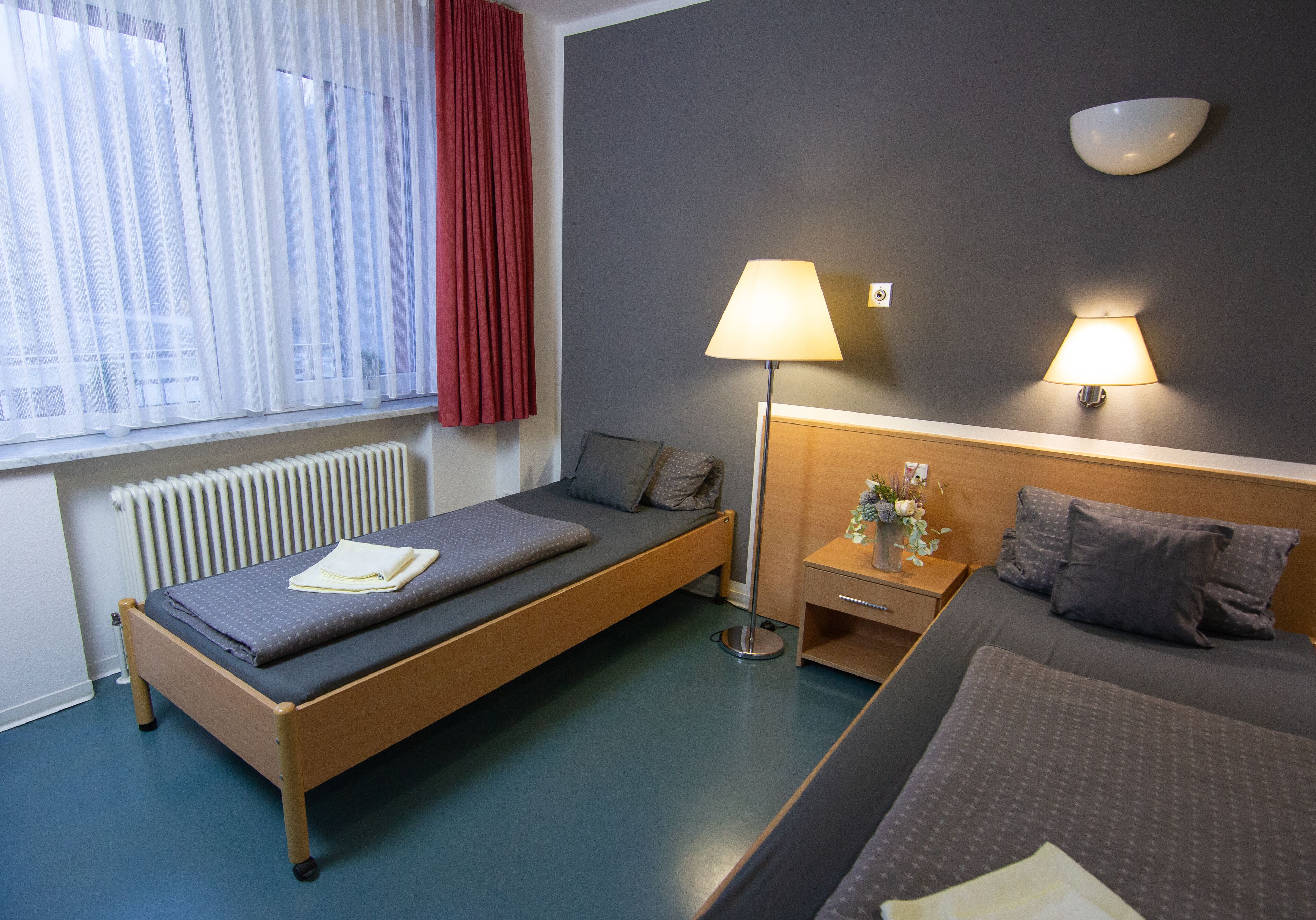 A hotel room with a single bed, a smaller bed, a bedside table with a lamp, a nightstand, a window with curtains, and a wall-mounted light fixture.