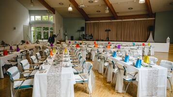 Une salle de banquet avec de longues tables dressées pour un événement formel, des poutres en bois au plafond et une scène avec des rideaux en arrière-plan.