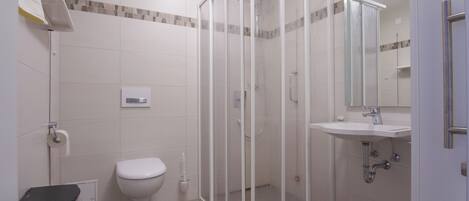 A bathroom with a glass shower enclosure, a toilet, a sink with a mirror, and a trash bin.