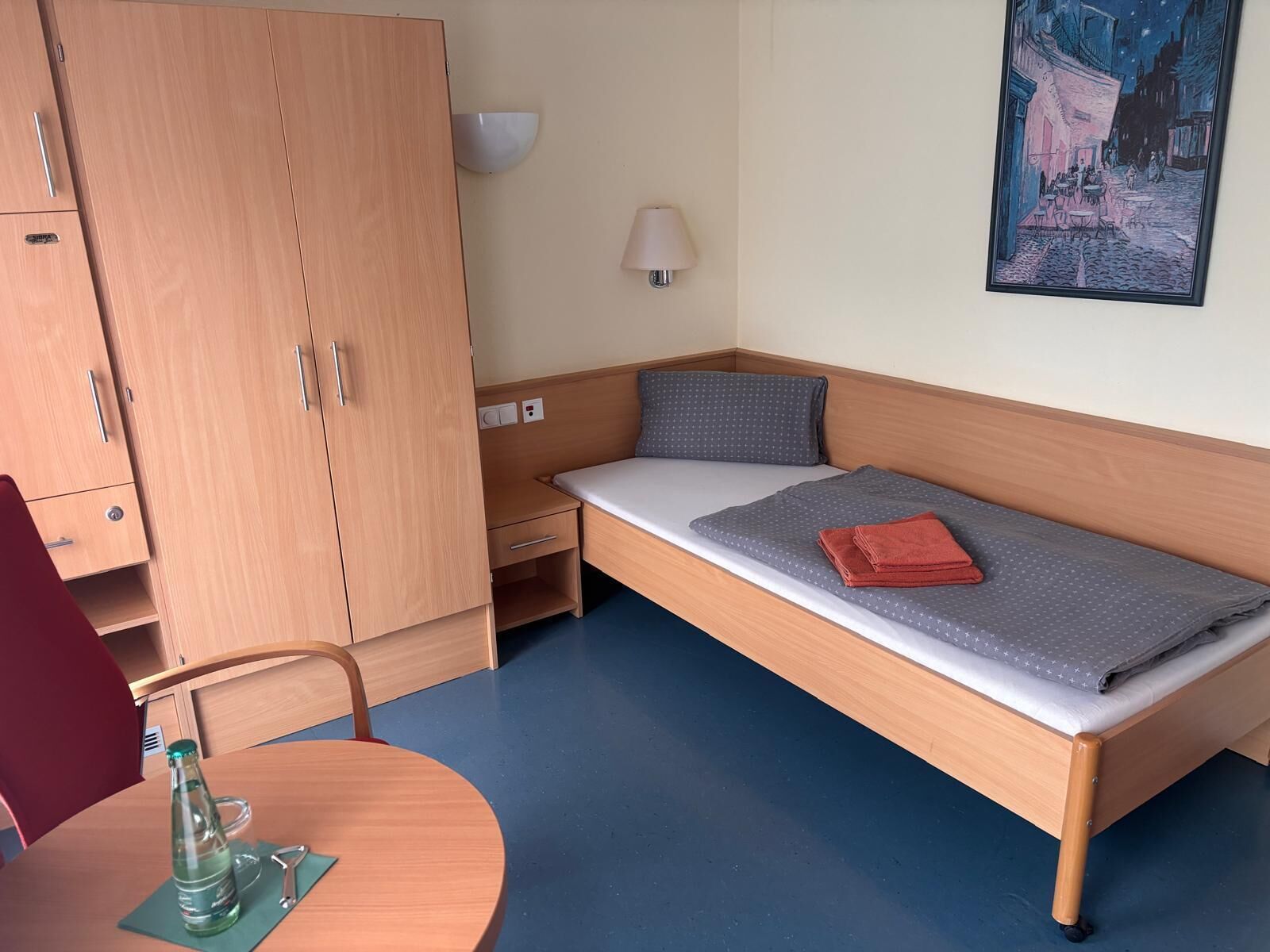 A single bed with a grey mattress and a red towel, a wooden wardrobe, a small bedside table, a lamp, and a painting on the wall.