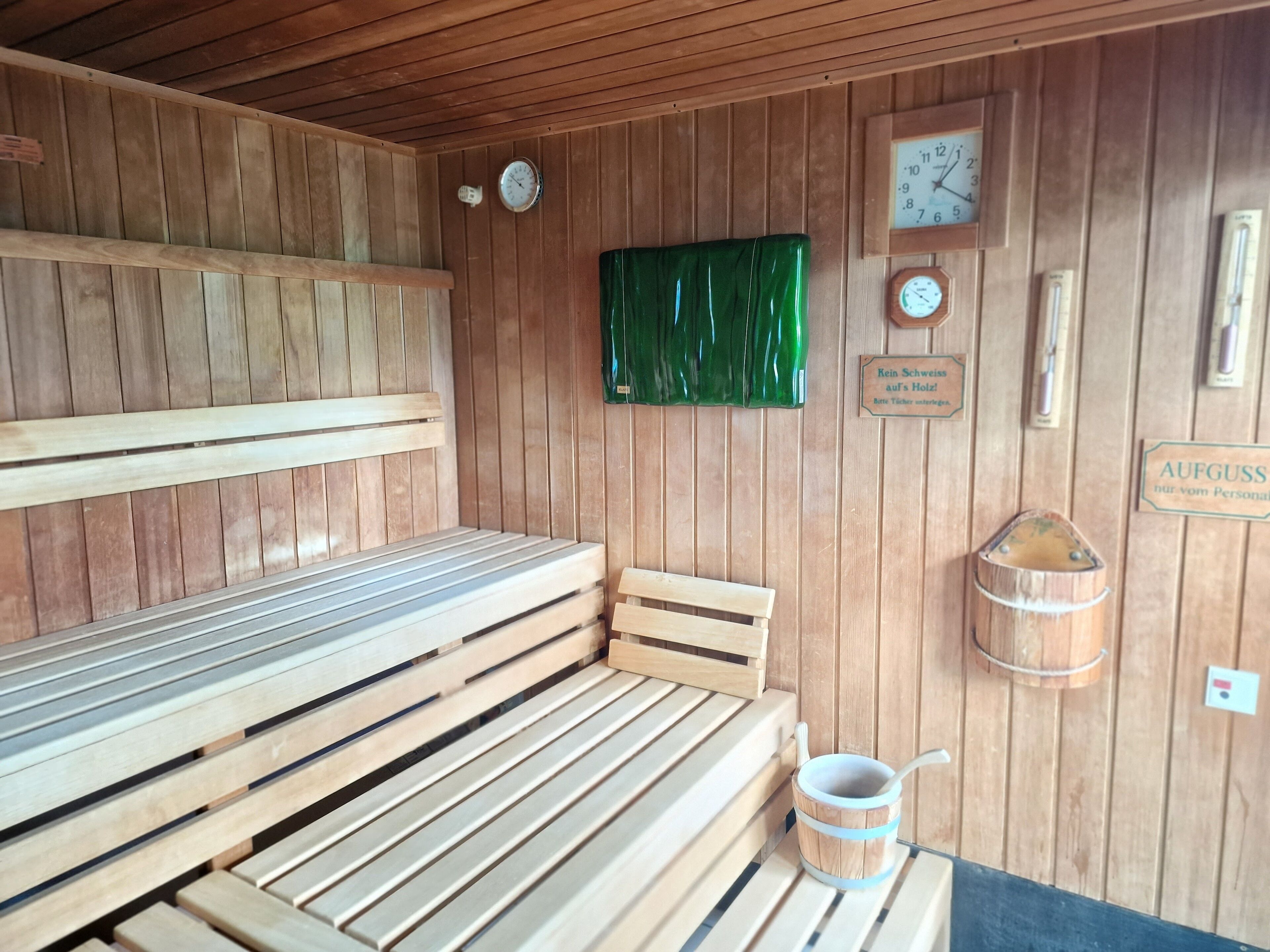 A wooden sauna with benches, a green curtain, a clock, and a sign in German.