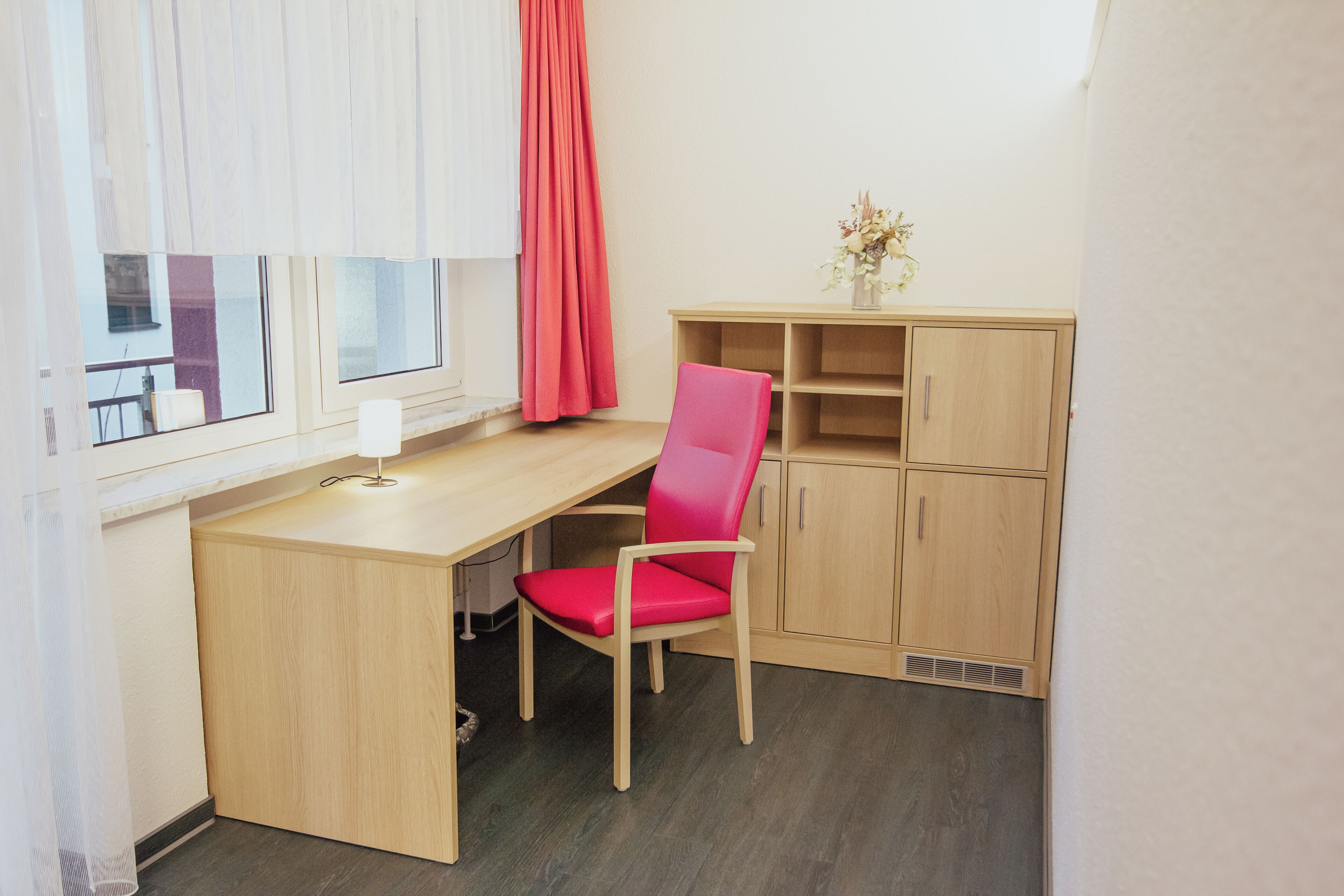 A small, compact room with a desk, a chair, a window with curtains, and a cabinet.