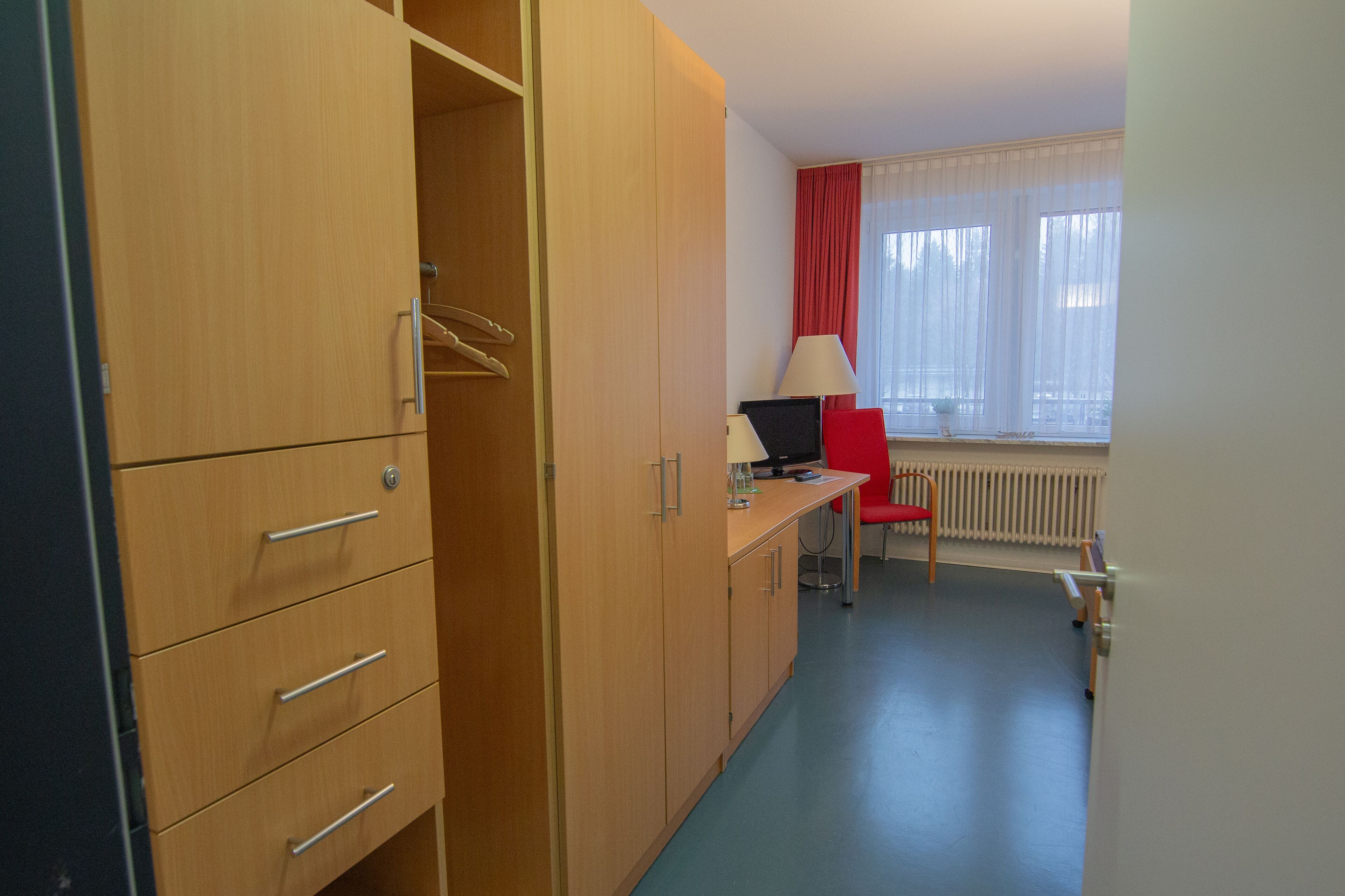 Una habitación de hotel con un armario de madera, un escritorio con lámpara, una silla roja y una ventana con cortinas.