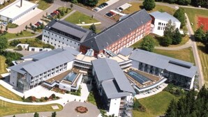 A modern building complex with multiple interconnected structures, surrounded by green fields and trees. - Green Night Hotel (Falkenstein/Vogtl.)