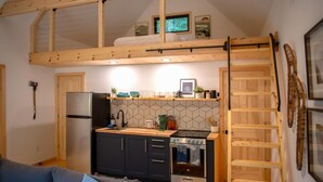 A compact kitchen with a built-in stove, a refrigerator, and a wooden loft above. - The Pines Cabin | New Tiny Home Near Mt Snow! (Wilmington)