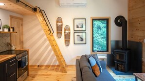 A cozy cabin interior with a wooden staircase, a sofa, a kitchen area, and a wood-burning stove. - The Pines Cabin | New Tiny Home Near Mt Snow! (Wilmington)