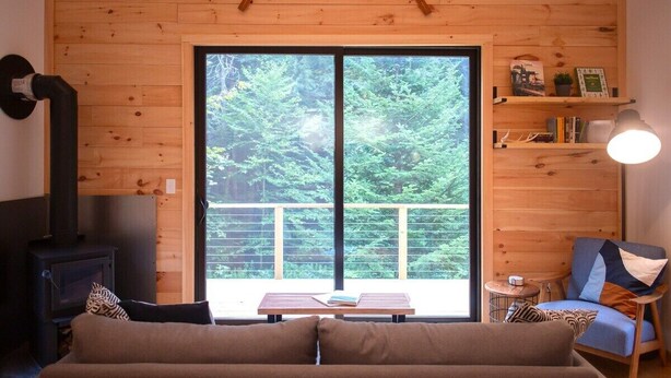 A cozy cabin interior with a wooden interior, a sofa, a table with books, a small table with a lamp, and a view of a forest through a large window. - The Pines Cabin | New Tiny Home Near Mt Snow! (Wilmington)